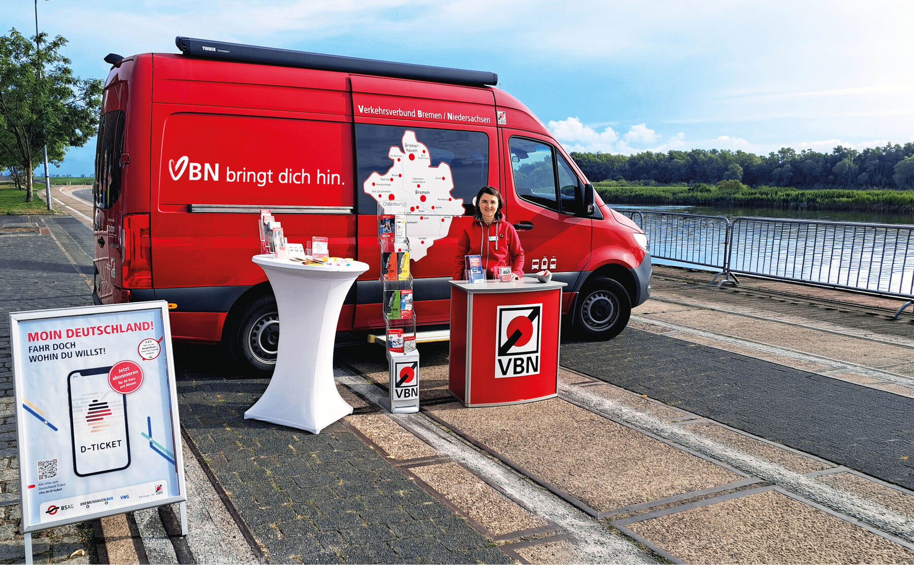 Rotes VBN-Infomobil mit Stehtisch, Infostand und Mitarbeiterin an einem Flussufer mit Blick auf Wasser und Bäume